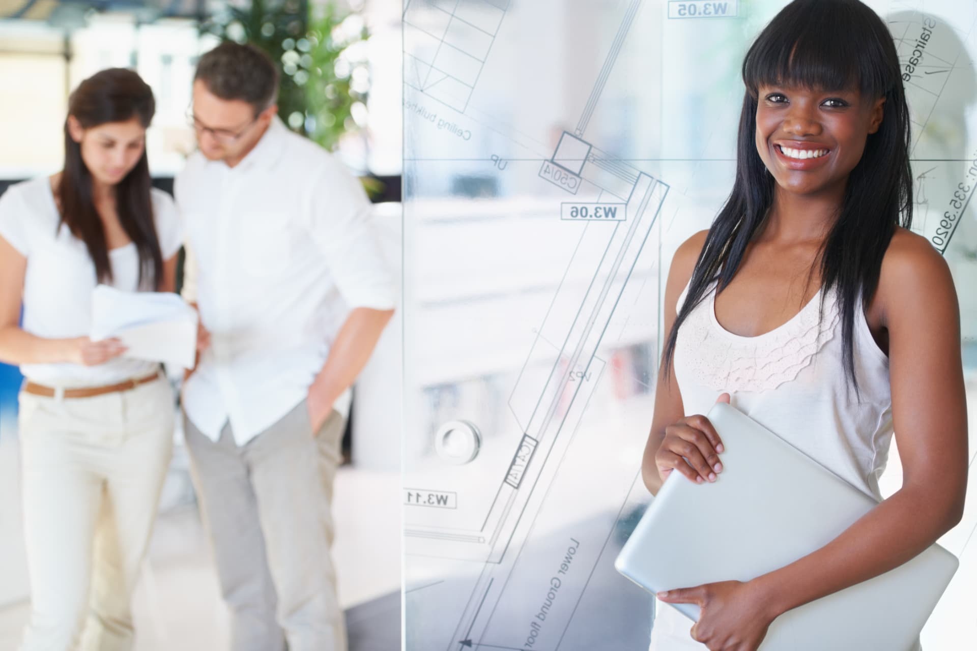 Smiling woman holding a laptop beside a glass wall printed with floor plans, two colleagues reviewing papers behind her.