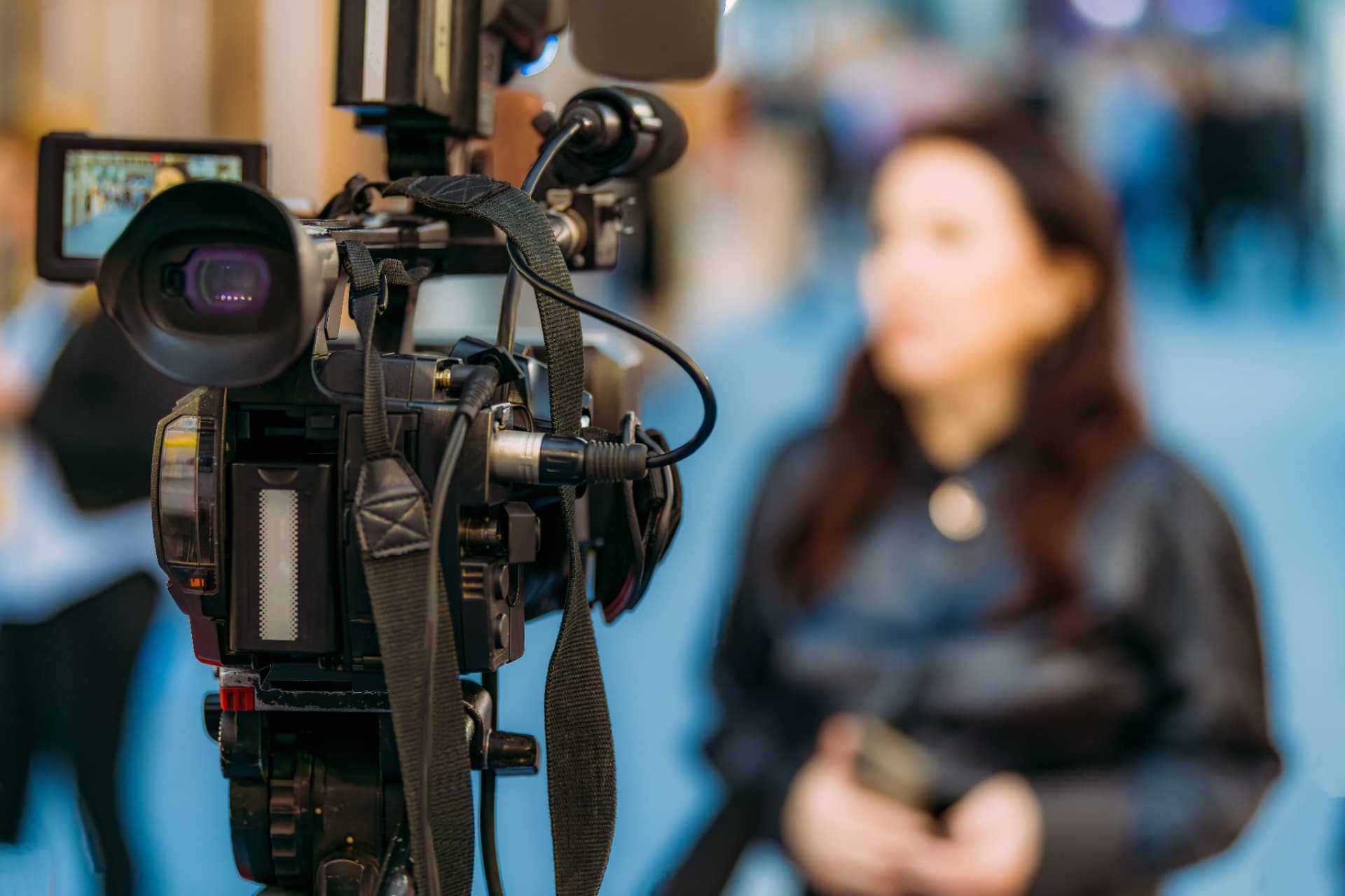Broadcast camera in sharp focus filming a woman being interviewed at a trade event.