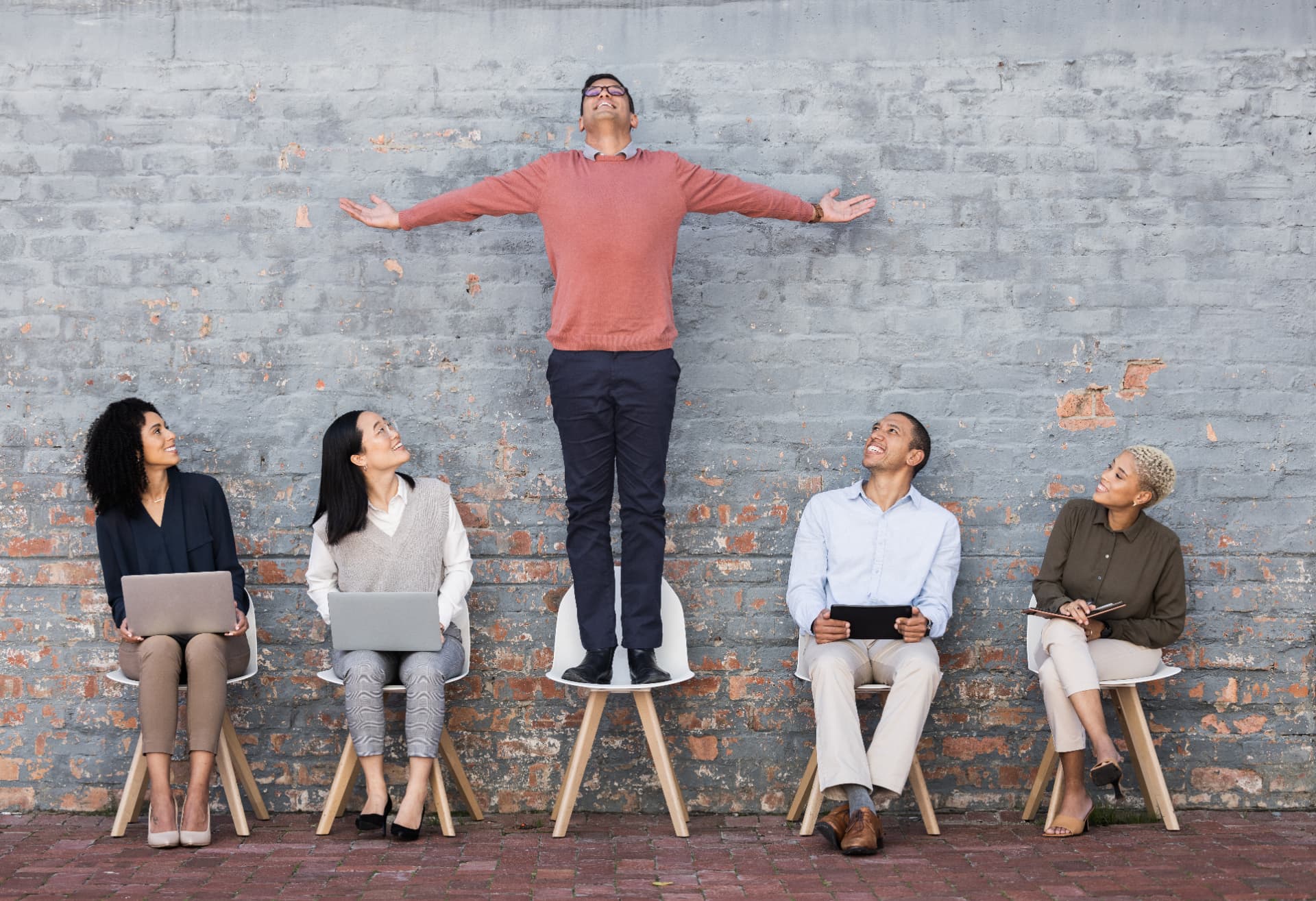 One man standing arms outstretched on a chair while four seated colleagues look up, against a grey brick wall.