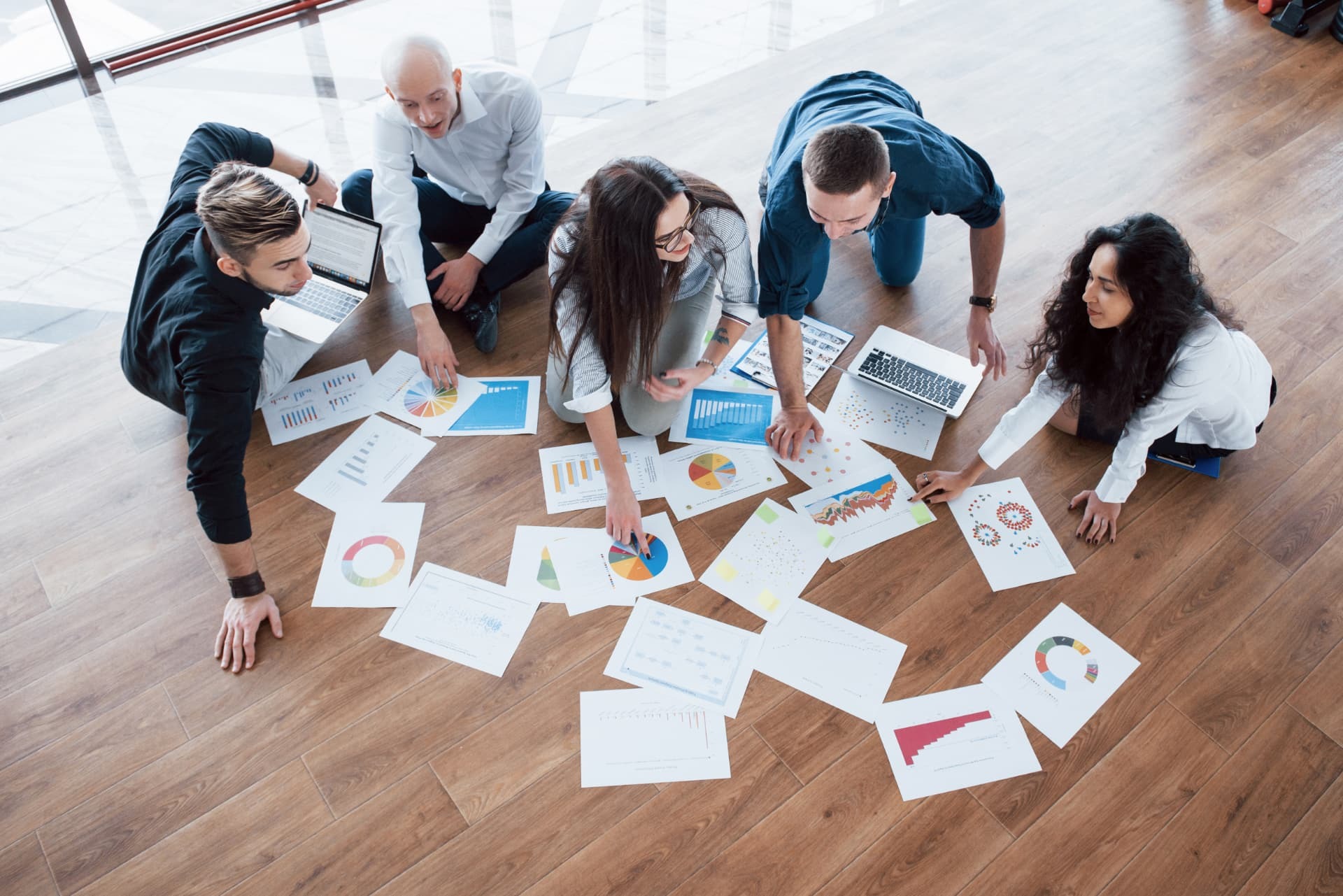 Five colleagues kneeling on a wooden floor around printed charts and laptops, planning together.